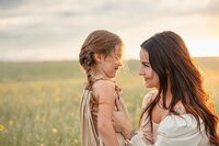A woman and a young girl with braided hair smile at each other in a sunlit field, captured beautifully by a Colorado Wedding Photographer. The woman holds the girls arms gently against a backdrop of a green meadow and cloudy sky.