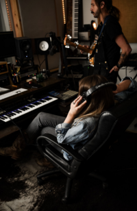 woman sitting in front of keyboard at music recording session with headphones on.  man standing  next to her