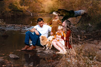 A family with a mom, dad, a young girl, a toddler, and a tan and white dog are sitting at the edge of a creek on rocks. They're looking at each othr and smiling. There's gorgeous fall yellow and orange leaves on the trees behind them.