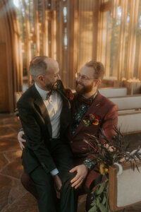 A man in a black suit sits in the lap of his just-married husband, whom is dressed in a dark red suit. They are sitting in the pews of Cooper Chapel in Bella Vista, Arkansas.