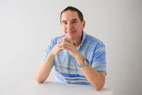 A professional headshot of a smiling man in a blue striped shirt, posing with hands clasped on a white background. Perfect for business and personal branding. Captured by Leslie Q Photography.