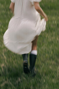 Woman walking through lush field in black boots and white dress
