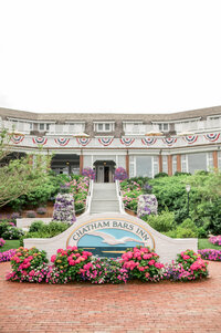 The welcoming entrance to Chatham Bars Inn, surrounded by lush gardens and blooming pink flowers, with a curved stairway leading up to the inn's iconic sign and grand façade.