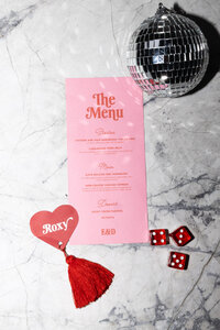 Pink, retro wedding menu and wedding guest name tag with a disco ball and dice next to it