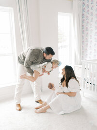 a mother and father sit in their newborn's nursery, the dad holds the big sister in the arm while mom holds the baby in her lap
