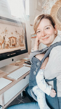 Jess Meyer at her studio desk with her baby in a carrier, her design work on the computer.
