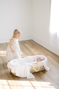 A big sister gently sneaks up on her new sister who is laying in a bassinet.