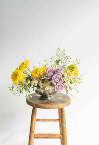Yellow and purple bouquet sits on wood stool in front of white wall