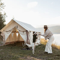 Sydney Breann, a Montana-based elopement photographer, plays with her dog beside a canvas tent near a mountain lake during a behind-the-scenes camping shoot.