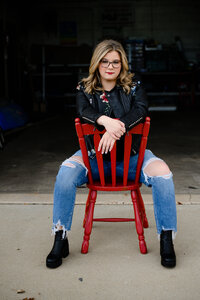 A denver senior photographer captures senior photos for a girl in a black leather jacket sitting backwards on a red chair for her denver senior pictures