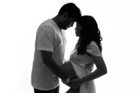 Couple posing for a maternity picture they are standing up nose to nose with hands on woman's pregnant belly. Woman is wearing a white sheer open maternity dress exposing her baby bump.