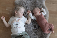 Livsstilsfoto av storesøster og lillesøster ligger på en grå ullfell og sover mens de holder hender.