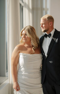 A woman smiles at the camera, looking over her shoulder. Behind her, slightly blurred, is her fiancé smiling big at her and wearing a suit.