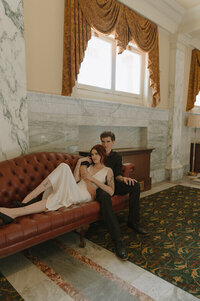 A couple sits on a red leather couch on their elopement day. He has his arm around her as she lays on his chest.