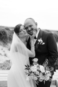 A groom smiles as his bride grabs his face for a kiss on a beach