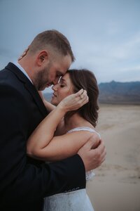 bride and groom hold each other as they stand on a sand dune with mountains behind.