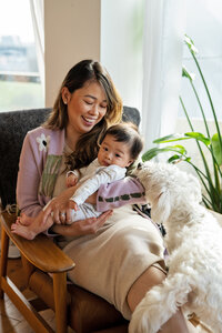 A mother sits and smiles as she cuddles with her baby boy. The family dog climbing up on a foot stool reaches to sniff at the baby as if he is kissing him.