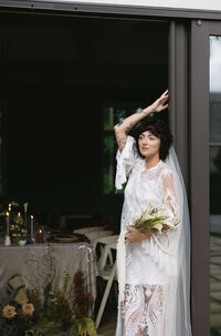 The bride poses near a door with a bouquet, wearing a lace dress