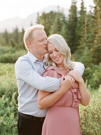 Couple hugging each other with the guy kissing the girls head and smiling together wearing a blush pink flowy engagement session dress and the guy wearing a light blue button up shirt standing in the middle of the rocky mountains with tall green trees in the background and photos by Tulsa Wedding Photographer Laura Eddy