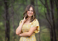 high school senior girl in a yellow dress playing with her hair standing amongst trees in Pueblo Colorado