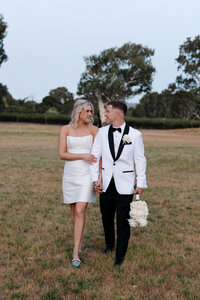 Bride holds grooms arm as they walk through a grass field.