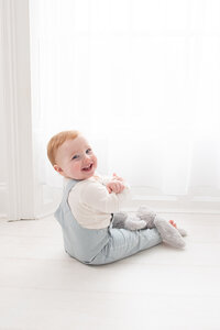 A baby with light hair is sitting on the floor by a bright window at Dandelion Studio in Moira, Northern Ireland. Dressed in a light blue outfit, the baby is turned slightly towards the window with a happy expression, creating a gentle and serene atmosphere.