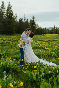 A man is kissing his fiance in a field with yellow wildflowers. She is wearing her wedding dress and he is wearing a button up with blue jeans.