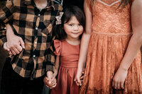 A warm moody portrait of a young girl in a deep coral dress with a soft smile standing between her older brother and sister holding their hands.  Their faces are not visible, only their hands and torsos can be seen. The older sister wears an orange lace dress and the brother wears a yellow and black shirt.  The shadows are rich and the light is soft and warm.