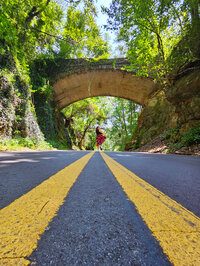 a girl in a colorful skirt standing in the middle of the road inder an overgrown bridge
