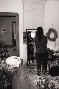 Black-and-white image of florist Roxy focused on designing an arrangement, surrounded by blooms in her studio.