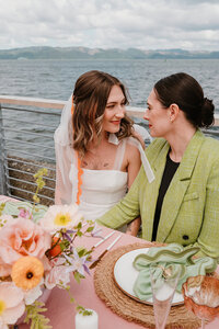 A wedding photo of two brides sitting at their sweetheart table. They are looking at each other with soft smiles. The woman on the left is wearing a white wedding dress with a veil that has orange wavy trim. She has soft brown hair with touches of blonde. The woman on the right is wearing a lime suit jacket with a black shirt. She has dark brown or black hair in an updo. The table setting is colorfully decorated with pink, green, peach, and lavender tones throughout. They are sitting near a railing that overlooks the Columbia River Gorge.