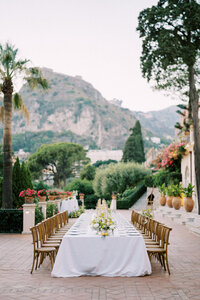 Elegant outdoor wedding reception table set for guests with floral centerpiece, surrounded by mountains and greenery in a romantic villa setting
