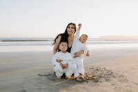 A mother with long dark hair smiles while holding her two young sons in white clothing on the beach at sunset.
