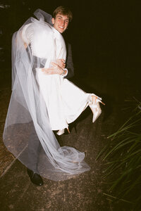 A grrom lifts his bride up in a fun and candid moment , her heels are muddy , and the feeling is vintage , retro and nostalgic