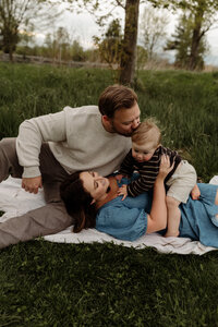 A family of three relaxes in a scenic field, captured beautifully by a New England photographer. The father gently kisses their baby’s head as the mother rests on his lap, cradling the little one on her tummy. Dressed in soft, timeless tones, they’re surrounded by lush greenery — a tender, fine art moment filled with warmth and connection.