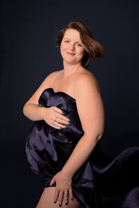 A person with a short hairstyle stands against a dark background, wrapped in a dark, flowing fabric that covers their body. They are smiling and looking towards the camera, capturing the essence of elegance posed by an award winning Jacksonville maternity photographer.