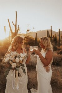 A couple toasts with glasses of champagne during their elopement at Sabino Canyon in Tucson, Arizona. Both are dressed in elegant white gowns, standing amidst a desert landscape at sunset, with cacti and mountains in the background.