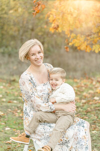 Mom in light colored floral dress holding son who is dressed in khakis and a cream sweater. They are both giggling and the sun is shining through the fall leaves behind them. Fall outdoor Portland Family Photography session.