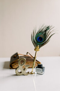 A journal, a feathered ink pen and bottle of ink, with a gold metal ampersand paperweight sitting on a white desk with a white background