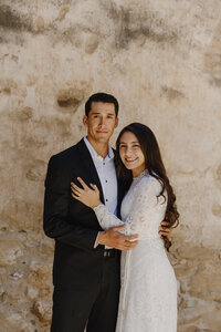 A couple standing close together smiling in front of a sunlit stone wall during their engagement session at Canopy Grove.