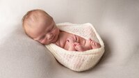 A newborn baby sleeps soundly, wrapped in a soft cream-coloured knitted blanket at Dandelion Studio in Moira, Northern Ireland. The baby is posed on a neutral-toned blanket, curled up in a cozy position with a peaceful expression, creating a sense of warmth and tenderness in this classic newborn portrait.