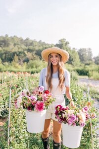 Flower farmer holding buckets of harvested flowers and smiling at camera with a hat on