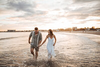 A couple at the beach running through the water at sunset holding hands.