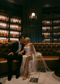 The groom and the bride are sitting on the sofa and kissing during a photoshoot at the hotel