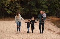 Family of 5 with 3 young children holding  hands and walking along the edge of a lake on the sandy beach