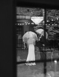 A Columbus wedding couple stands outdoors under clear umbrellas on a rainy day, sharing vows. They're framed by a window, with string lights and a tree in the background.