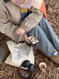 a woman sitting on a dried pile of leaves holding her phone, headphones, and coffee