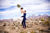 Groom lifts bride in celebration atop a rugged mountaintop at Grand Targhee Resort, with the Grand Tetons stretching across the horizon under a bright blue sky.