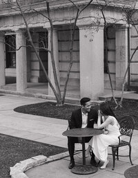 Black and white wedding photo of a smiling couple seated at a round metal table, surrounded by leafless trees and a classical building facade, conveying romance.
