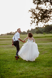 A couple running in a meadow. The groom is wearing suspenders, and is looking back as the couple runs